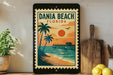 A framed poster of a tropical beach scene with palm trees, a pier, and a sunset over the ocean, displayed on a shelf next to a wooden cutting board and a potted plant.