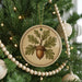 A round ornament with a tree and an acorn is hanging on a Christmas tree, surrounded by other ornaments and lights.