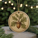 A round ornament featuring an acorn and leaves, hanging from a string against a blurred background of a Christmas tree with lights.