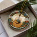 A decorative plate with a guitar design is placed inside a box, surrounded by pine branches.