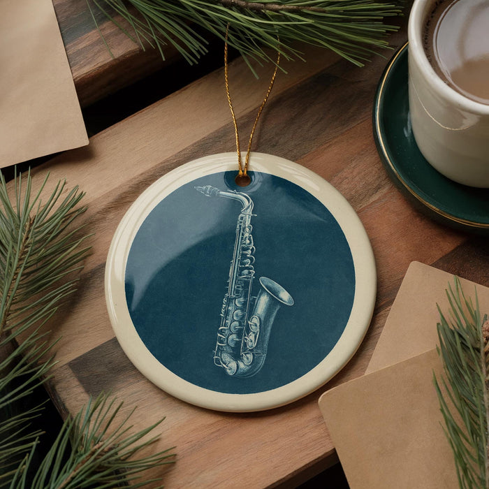 A blue ceramic ornament with a white rim, featuring a saxophone design, is displayed on a wooden surface alongside a white mug and some pine branches.