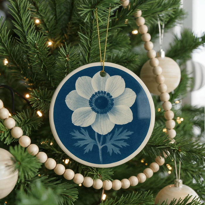 A blue and white circular ornament with a white flower design hangs from a Christmas tree, surrounded by white wooden beads and twinkling lights.