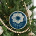 A blue and white circular ornament with a white flower design hangs from a Christmas tree, surrounded by white wooden beads and twinkling lights.