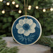 A blue and white ceramic ornament featuring a white flower with a blue center is displayed on a table, surrounded by pine branches and twinkling lights.