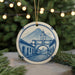 A decorative ceramic ornament featuring a detailed illustration of a town with a clock tower, set against a backdrop of a mountain and a bridge.