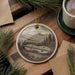 a round ornament with a black and white illustration of a lute, hanging on a wooden surface. The ornament is surrounded by pine branches and a cup of coffee, creating a cozy and artistic atmosphere.