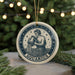 A circular ornament featuring the Aquarius zodiac sign, with a figure of a man pouring water from a pitcher. The ornament is hanging from a string and is surrounded by pine branches and twinkling lights, creating a festive atmosphere.