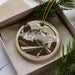 A circular ornament with a graphic design depicting a plane and mountains, placed in a box with pine branches.