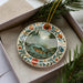 A decorative plate with a painting of a fish in a river, surrounded by a floral border, is displayed in a box with pine branches.