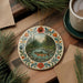 A decorative plate with a painting of a fish in a river, surrounded by a floral border, is placed on a wooden surface next to a cup of coffee and some pine branches.