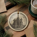 A decorative ornament featuring a detailed illustration of a cathedral or church, with multiple domes and spires, is displayed on a wooden surface alongside a cup of coffee and some pine branches.