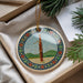 A decorative ornament featuring a musical instrument, specifically a clarinet, is displayed in a box with pine branches and a white card.
