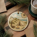 A round ornament depicting a camping scene with a tent, campfire, and pine trees, along with a compass rose, is displayed on a wooden surface.