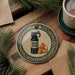 A ceramic ornament with a bear spray bottle and whistle on a wooden surface, surrounded by pine branches and a cup of coffee.