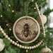 A round ornament featuring a bee is hanging on a Christmas tree, surrounded by other ornaments and lights.