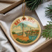 A decorative ornament with a circular design featuring a monument and a river, surrounded by greenery.