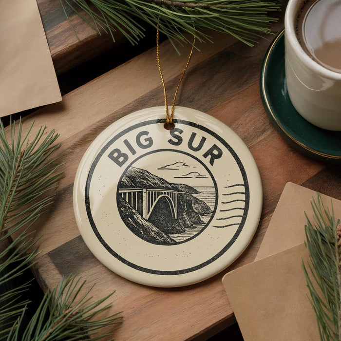 A decorative ornament with the text "BIG SUR" and an illustration of a bridge is displayed on a wooden surface, surrounded by pine branches and a cup of coffee.