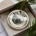 A decorative ornament in a box, featuring a black and white illustration of a bridge and the text "BIG SUR" on a circular design.