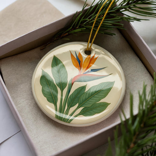 A decorative ornament with a floral design, featuring a large orange flower surrounded by green leaves, is displayed in a box with pine branches.