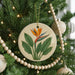 A round ornament with a tropical flower design hangs from a Christmas tree, surrounded by white beads and greenery.