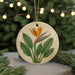 A round ceramic ornament with a floral design featuring a large orange flower surrounded by green leaves, placed on a surface with pine branches in the background.