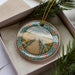 A decorative ornament in a box, featuring a wooden bridge and a scenic landscape.