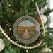 A Christmas tree decorated with ornaments, including a circular ornament featuring a wooden bridge and a field.
