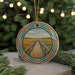A round ornament featuring a wooden bridge over a river, surrounded by a colorful border and set against a blurred background of greenery and twinkling lights.