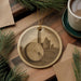a round ornament with a picture of a castle on it, placed on a wooden surface next to a cup of coffee and some pine branches.