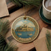 A decorative ornament featuring a castle and boats, with the word "BODrum" written on it, is displayed on a wooden surface alongside a cup of coffee and some pine branches.