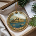 A decorative ornament featuring a castle and boats, with the word "BODRUM" written on it, is displayed in a box with pine branches.