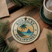A round ornament with the text "BORACAY PHILIPPINES" and a palm tree illustration, placed on a wooden surface next to a cup of coffee and some pine branches.