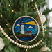 A stained glass ornament featuring a lighthouse is hanging on a Christmas tree, surrounded by ornaments and lights.