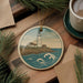 A round ornament featuring a lighthouse on a cliff overlooking the ocean, surrounded by pine branches and a cup of coffee on a wooden surface.