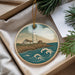 A decorative ornament featuring a lighthouse on a cliff overlooking the ocean, surrounded by pine branches.