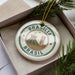 A circular ornament with the text "BRASIL" and an image of the Christ Church Cathedral in Brasilia, Brazil, is displayed in a box with pine branches.