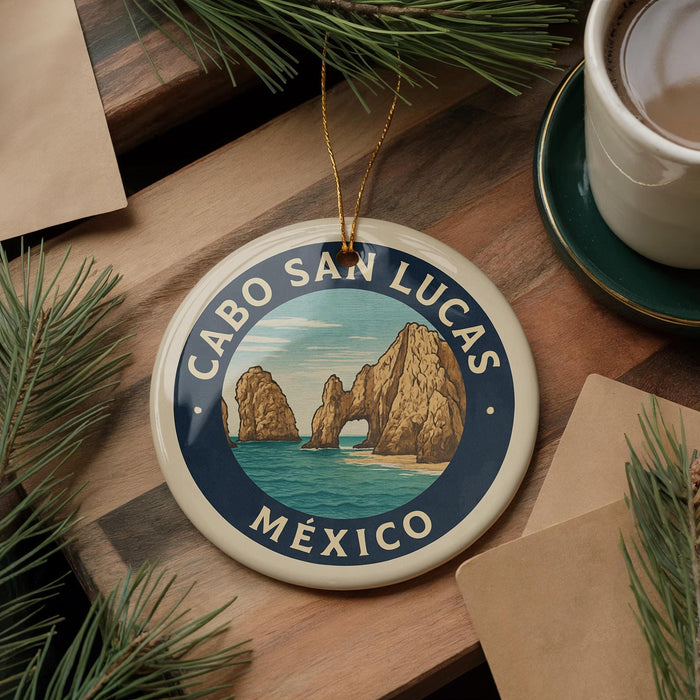 A decorative ornament featuring a scenic image of Cabo San Lucas, a popular tourist destination in Mexico. The ornament is placed on a wooden surface, surrounded by pine branches and a cup of coffee.