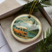 A round ornament featuring an orange van on a road, surrounded by pine trees and mountains, with a scenic landscape in the background.
