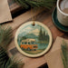 A round ornament featuring an illustration of a camper van on a road, surrounded by pine trees and mountains, with a lake in the background.
