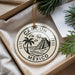 A circular ornament with the text "CANCUN MEXICO" printed on it, surrounded by a palm tree and a pyramid, is displayed in a box with pine branches.