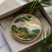 A round ornament with a painting of a person carrying a canoe on a river, surrounded by pine trees and a scenic landscape.