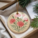 A round ornament with a floral design featuring pink carnations is displayed in a box, surrounded by pine branches and greenery.