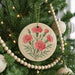 A round ornament with a floral design hangs from a Christmas tree, surrounded by other ornaments and lights.