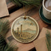 A decorative ornament featuring the city of Carthago, with a scenic illustration of the city's landmarks and a banner that reads "Carthago". The ornament is hanging from a string, and there is a cup of coffee and some pine branches in the background.