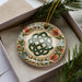 A decorative ceramic ornament with a Celtic knot design is displayed in a box, surrounded by pine branches.