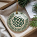 A decorative ornament with a green and white design featuring a Celtic knot pattern is displayed in a box, surrounded by pine branches and a white card.