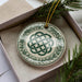 A decorative ornament in a box, featuring a circular design with a green and white color scheme and a Celtic knot pattern.