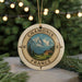 A decorative ornament with a compass design, featuring a mountain landscape and a small cabin, is displayed in front of a Christmas tree with twinkling lights.