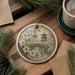 A decorative ornament featuring a Japanese temple and cherry blossom flowers, hanging on a wooden surface alongside a cup of coffee and some pine branches.