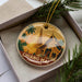 A decorative ornament featuring a temple with a golden spire and palm trees, placed in a box with pine branches.